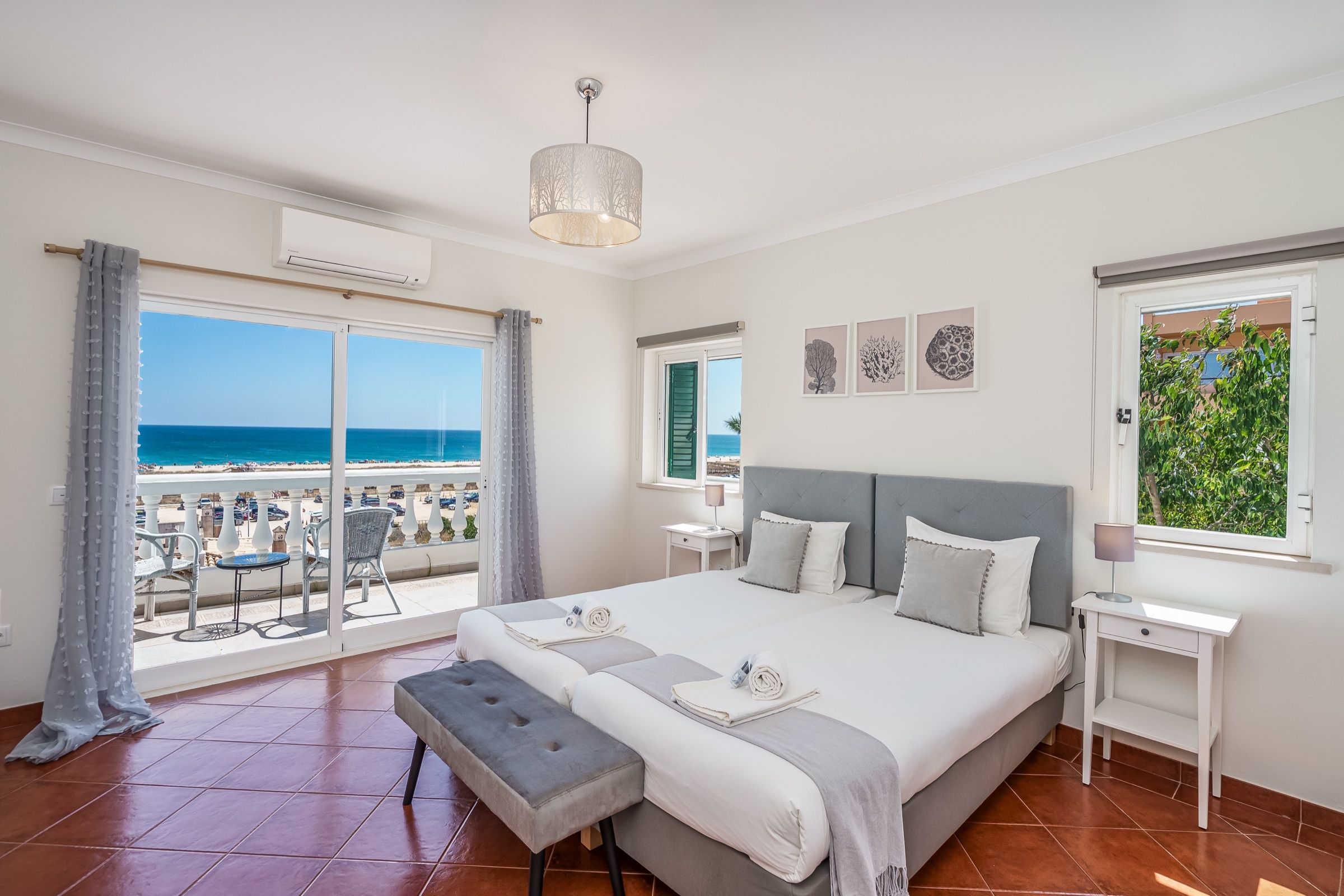 Quarto de casal com cabeceira cinzenta e porta envidraçada para a varanda, com o mar ao fundo