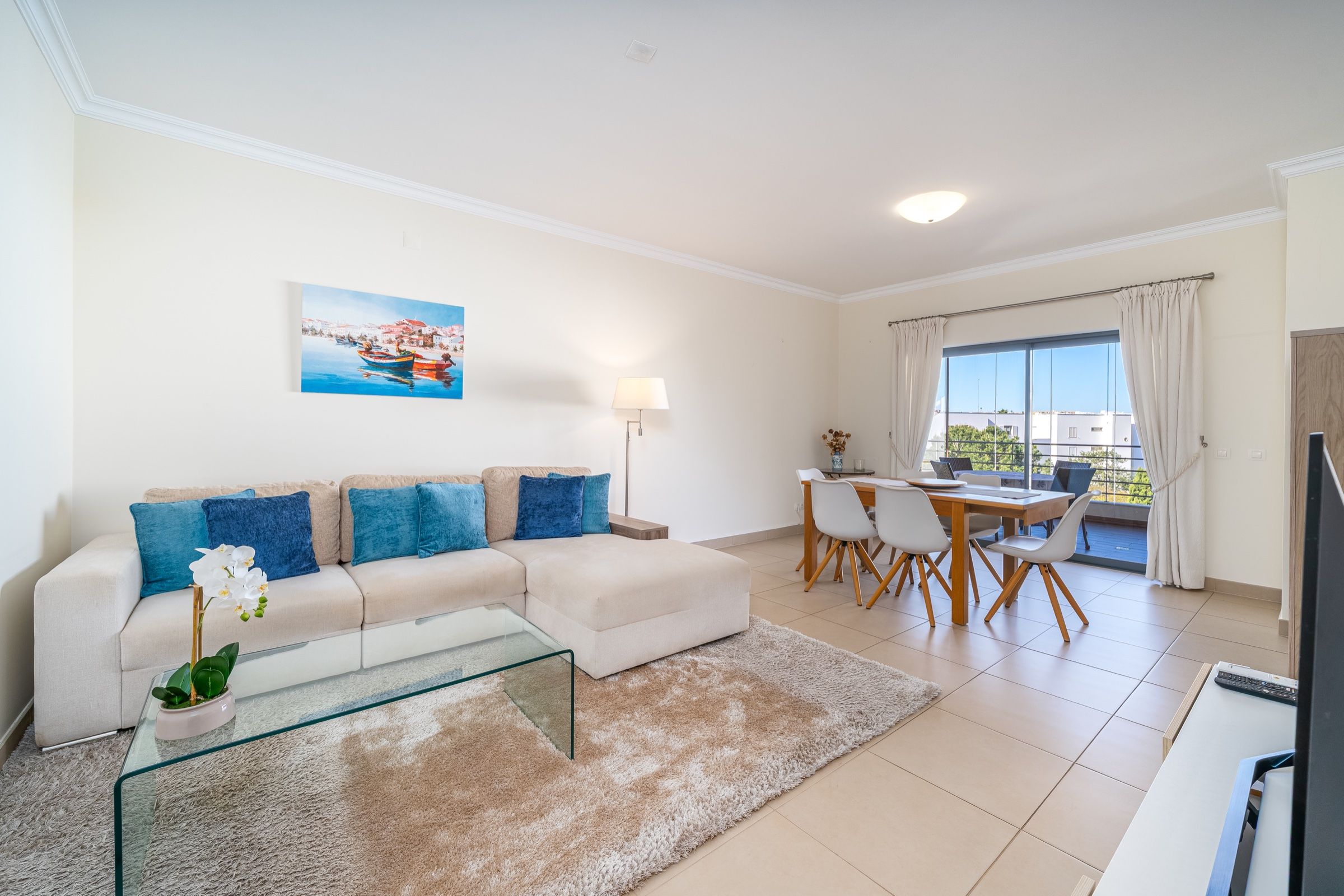 Sala clara com sofá de canto branco, almofadas azuis e mesa de jantar junto à janela
