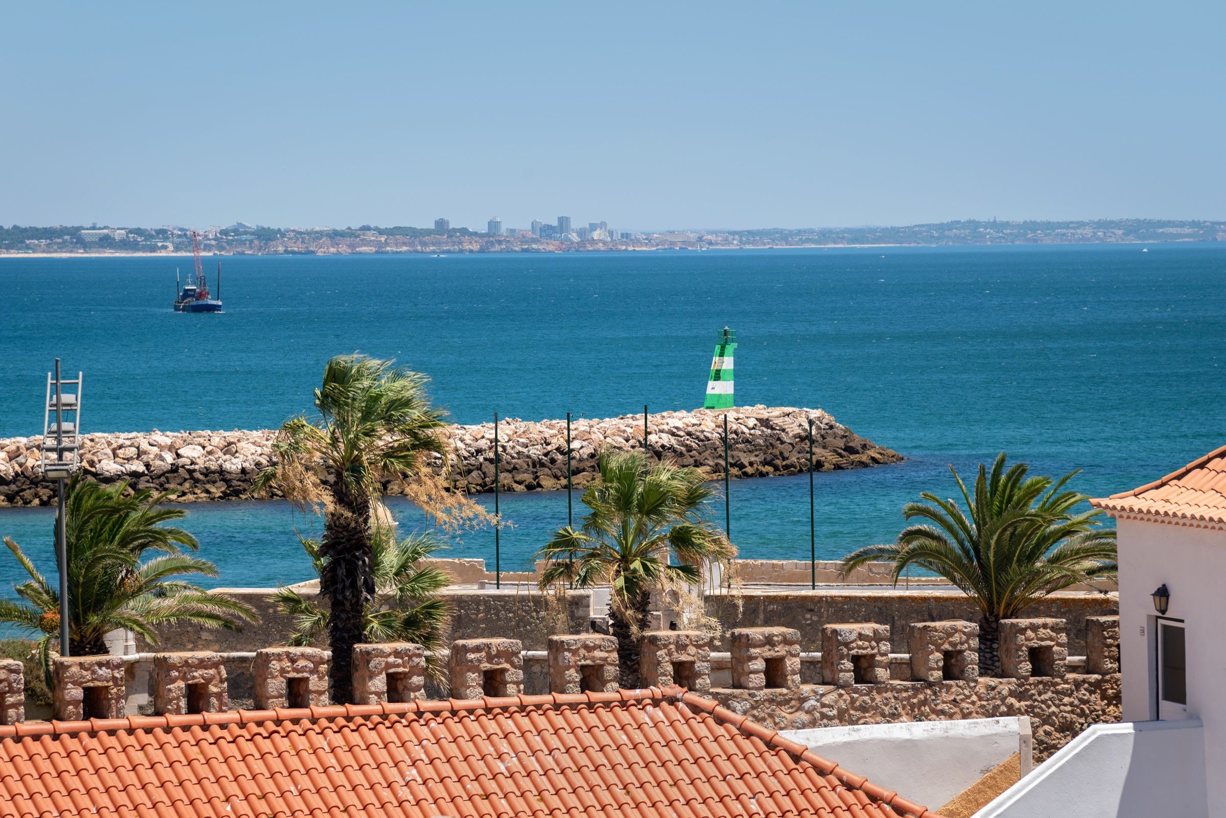 Vista sobre telhados e as muralhas da cidade para o mar, com o farol verde do molhe e palmeiras