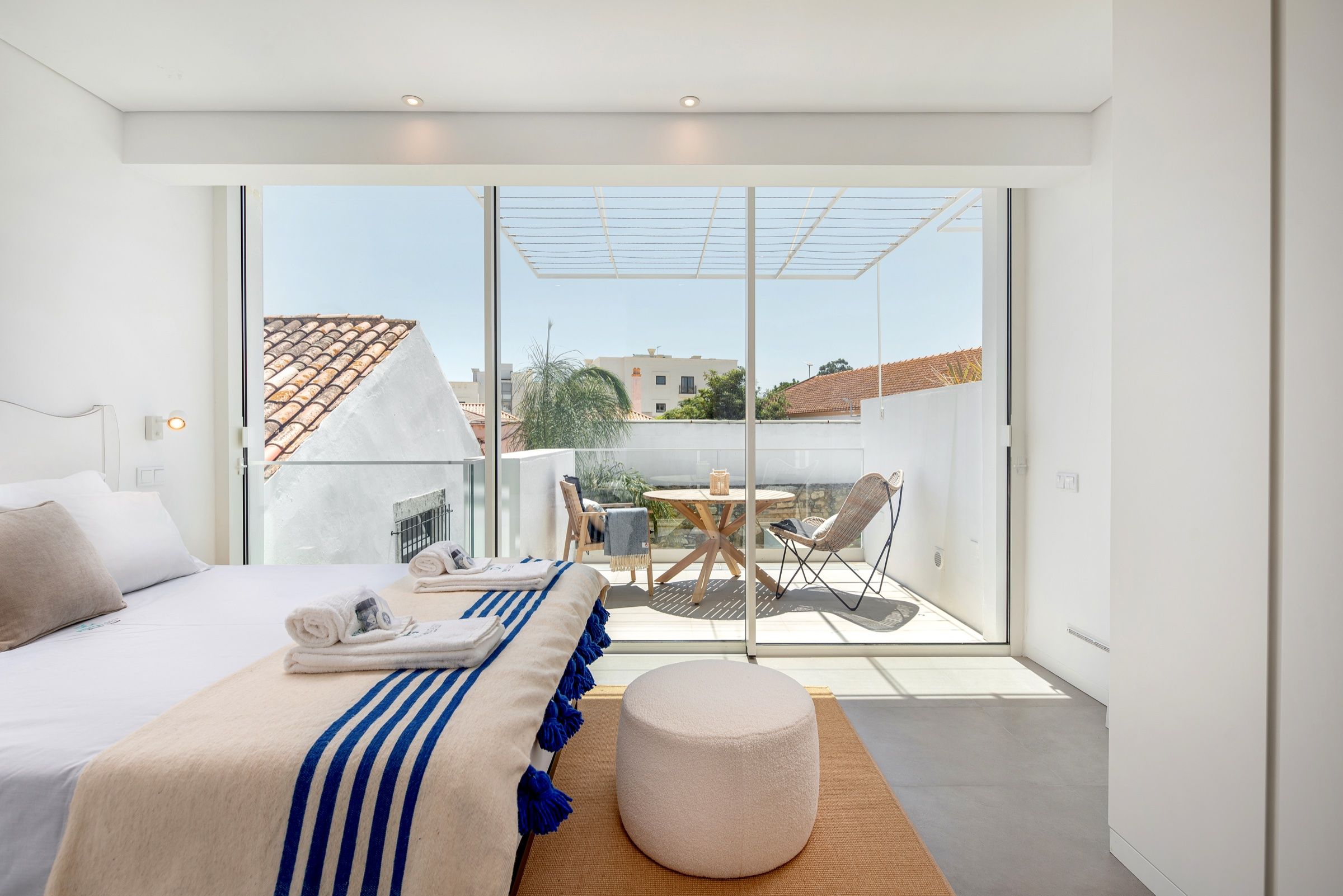 Quarto branco com manta às riscas azuis e porta envidraçada para um terraço com mesa e cadeiras