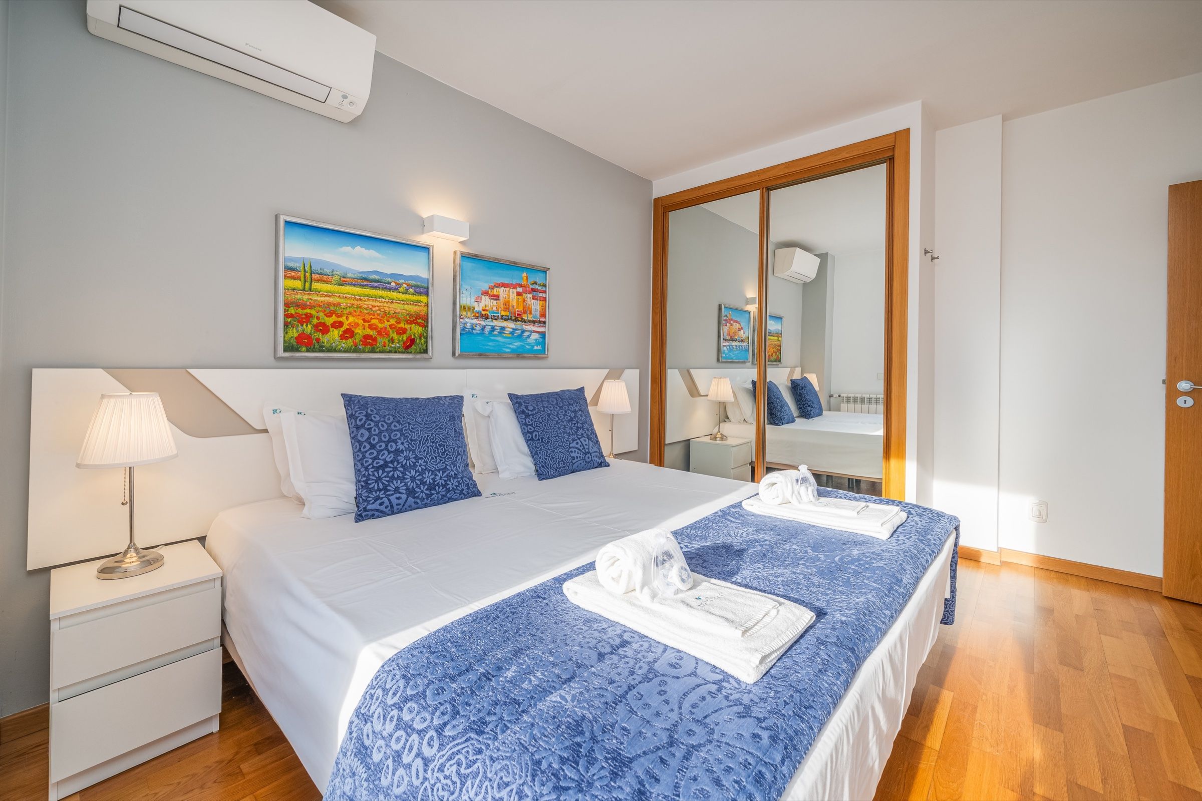 Quarto de casal com colcha azul, toalhas dobradas na cama e roupeiro com espelho