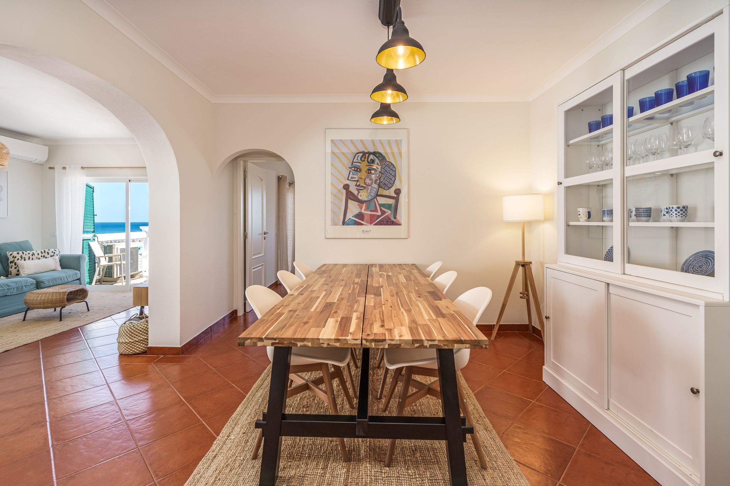 Sala de jantar com mesa comprida de madeira, cadeiras brancas, arcos e um armário com louça