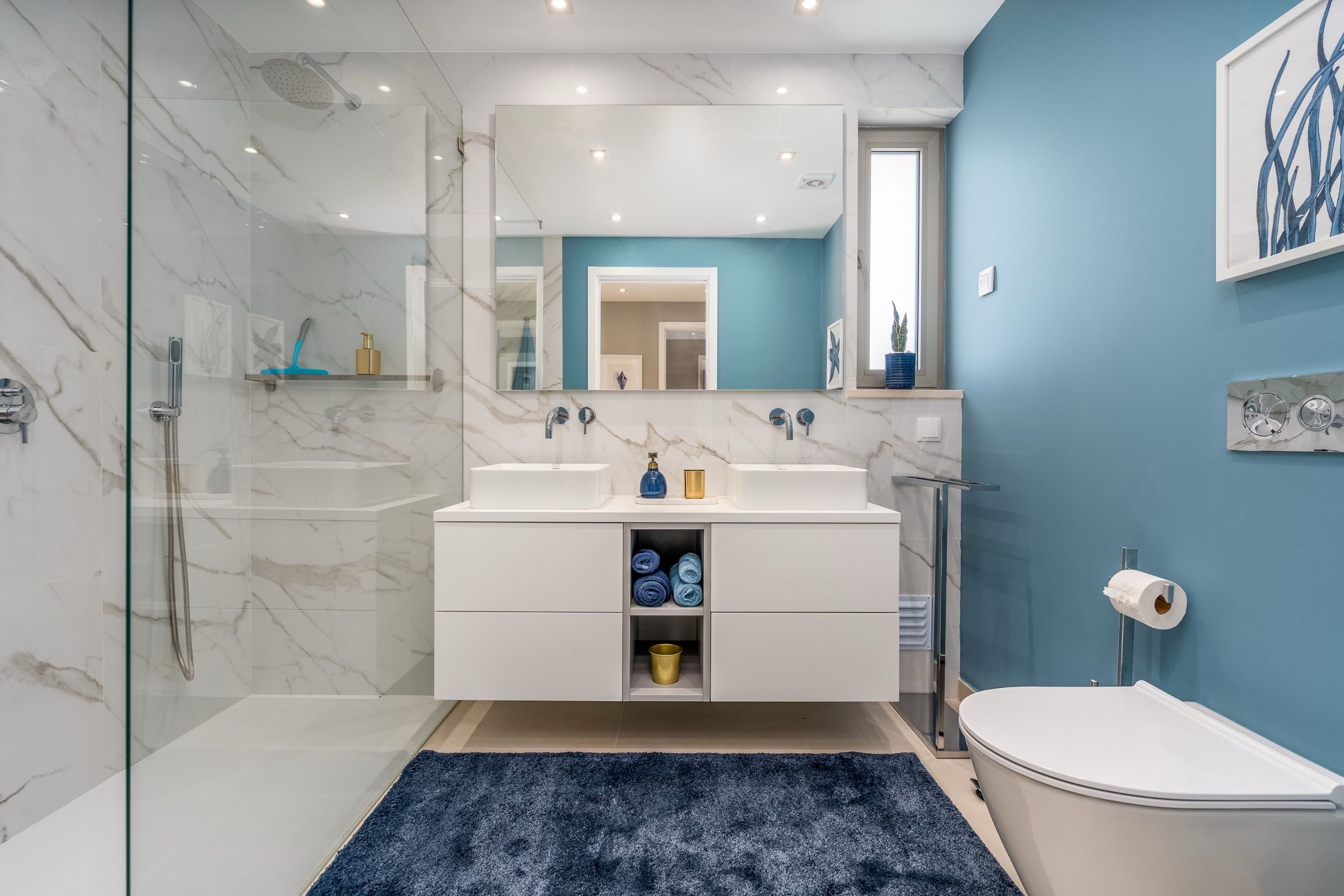 Casa de banho em mármore branco e parede azul, com duche de vidro e dois lavatórios