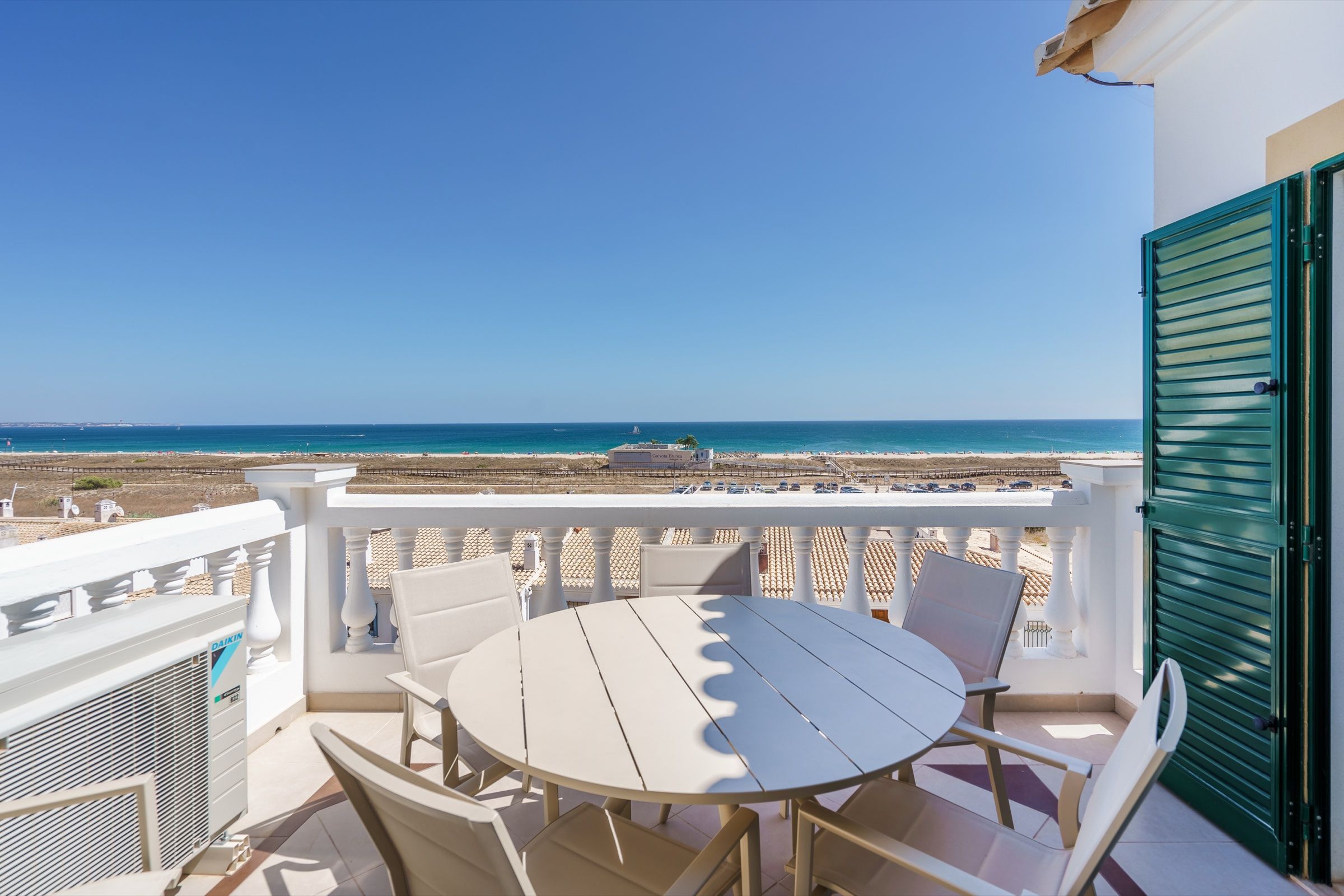 Varanda com mesa redonda e cadeiras, balaustrada branca e portadas verdes, virada para a praia e o mar
