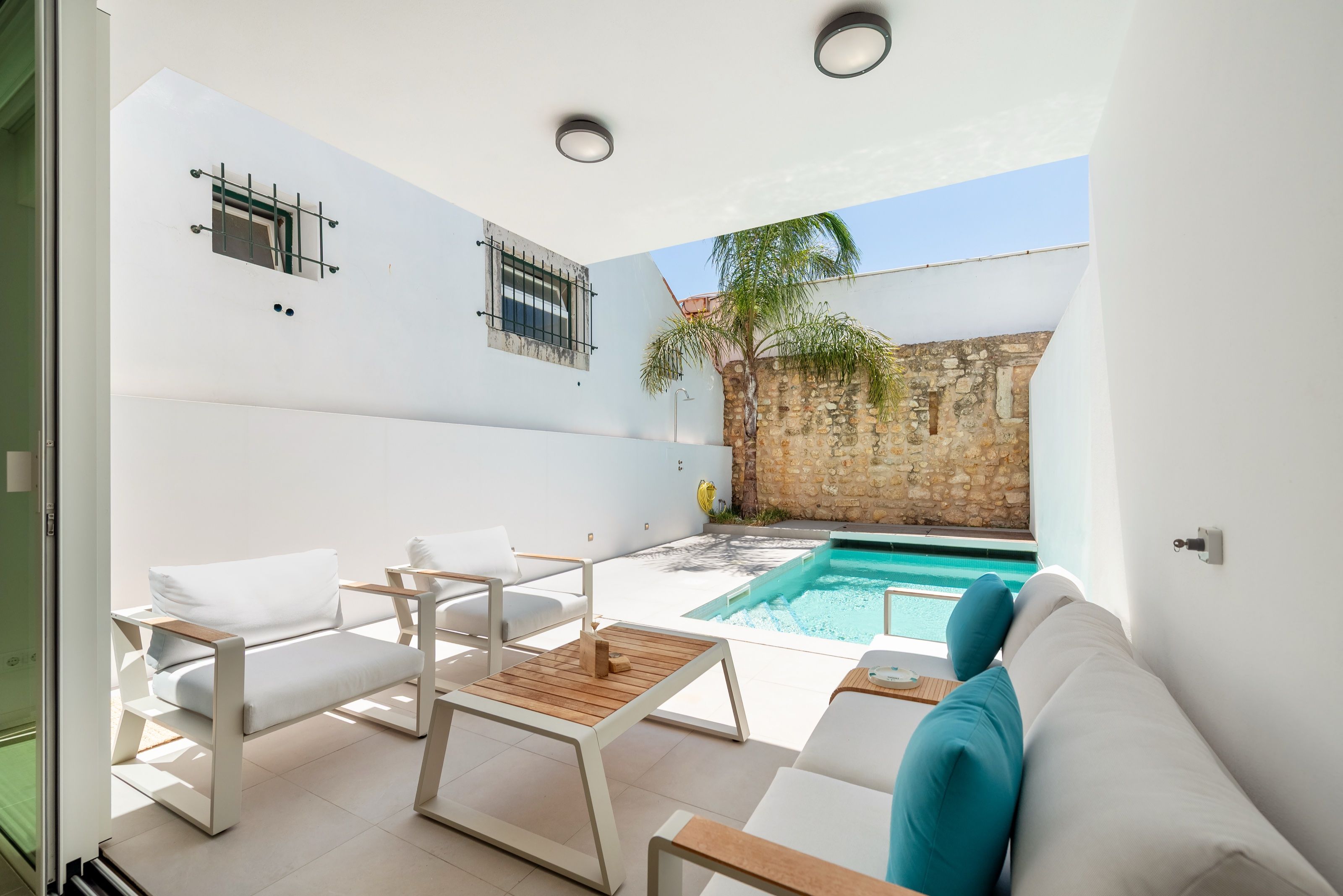 Pátio coberto de uma casa branca com sofás de exterior, a abrir para uma piscina com uma palmeira e um muro de pedra