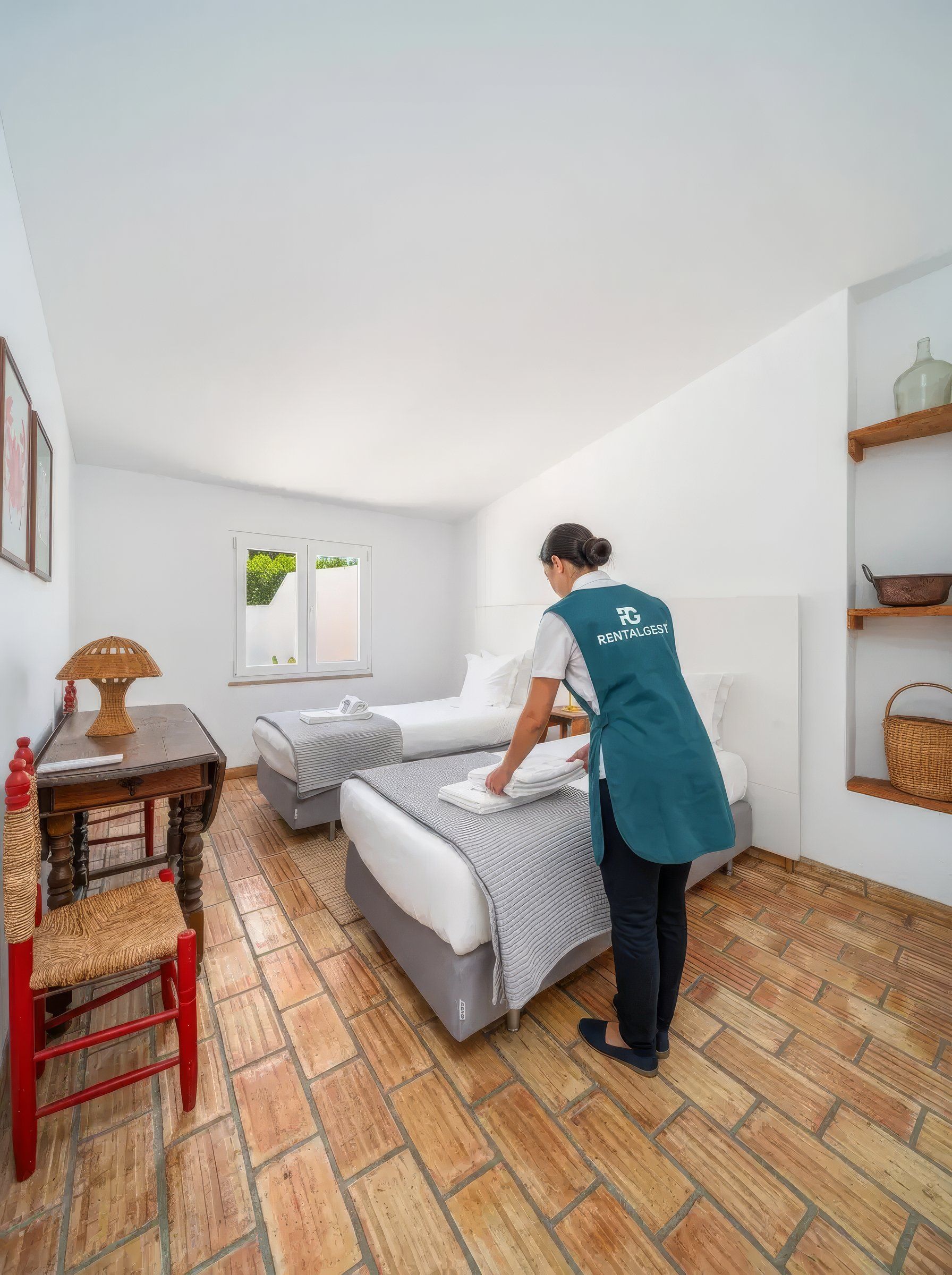 Funcionária com avental da Rentalgest a pôr toalhas dobradas numa das camas de um quarto com duas camas de solteiro e chão de tijoleira